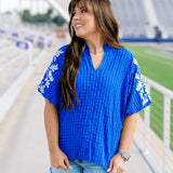 Woman with dark hair standing in front profile position wearing the Havanah Top at a football field. The focal point of this image is the intricate fabric on the front of the top, the embroidery on sleeves, ruffled collar, and v-neckline. The model paired this top with light-wash denim shorts.
