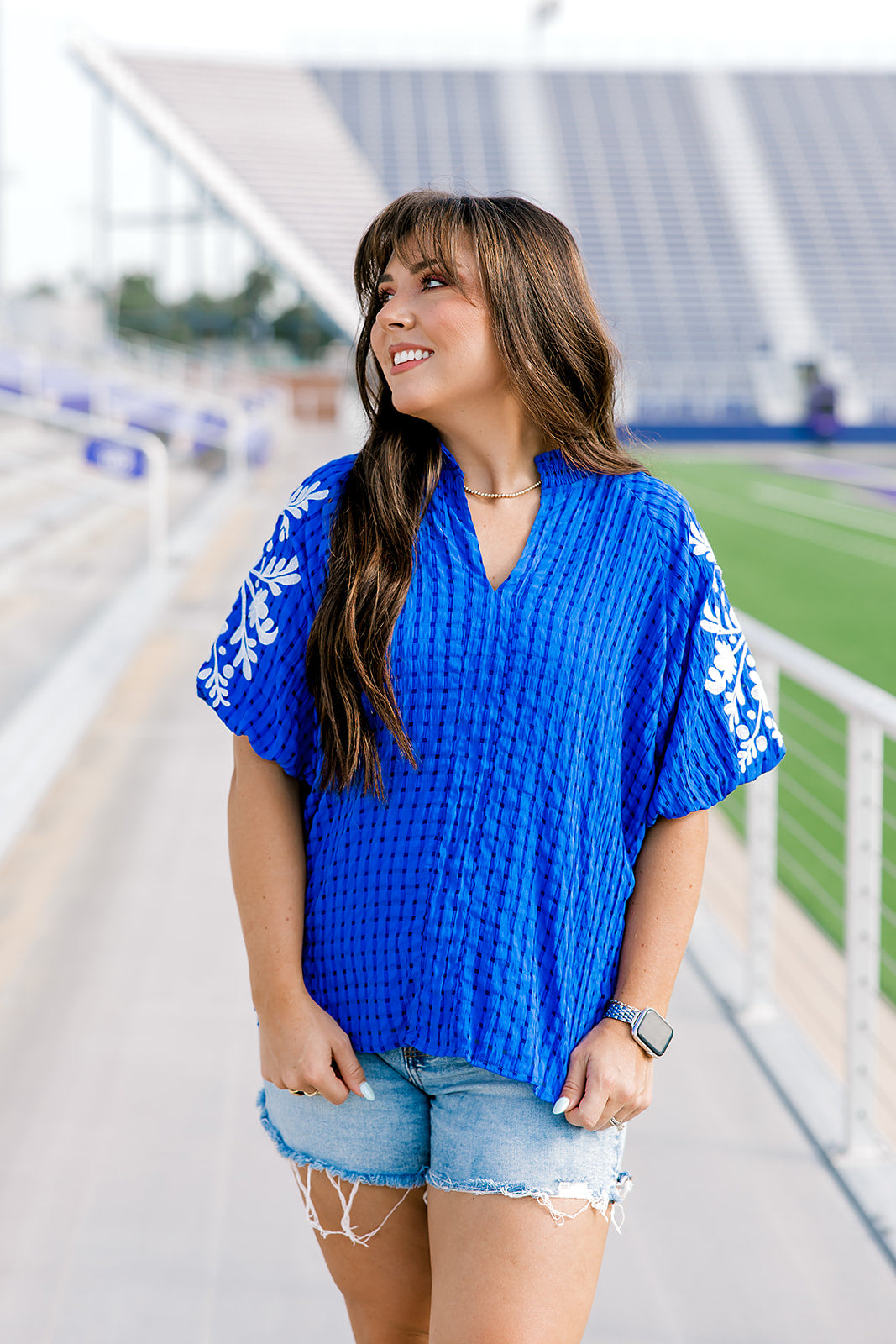 Woman with dark hair standing in front profile position wearing the Havanah Top at a football field. The focal point of this image is the intricate fabric on the front of the top, the embroidery on sleeves, ruffled collar, and v-neckline. The model paired this top with light-wash denim shorts.