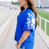 Woman with dark hair standing in a side profile position wearing the Havanah Top at a football field. The focal point of this image is the ruffled sleeves with embroidery over the shoulders, still showing the intricate fabric details. The model paired this top with light-wash denim shorts.