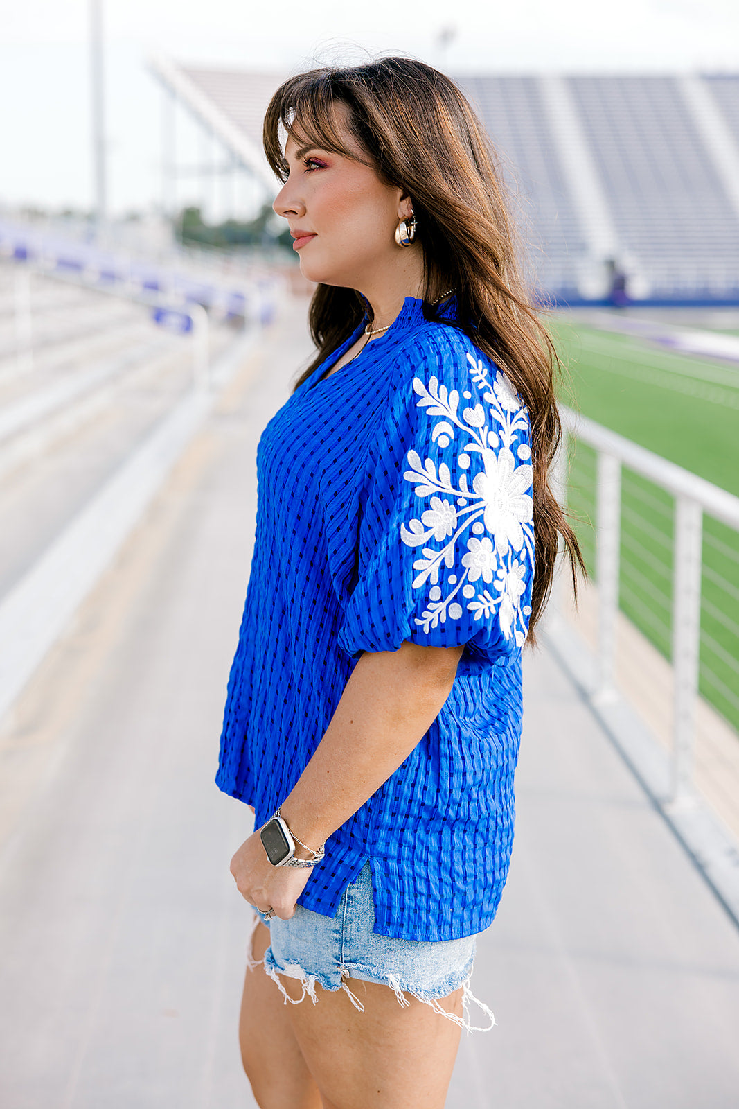 Woman with dark hair standing in a side profile position wearing the Havanah Top at a football field. The focal point of this image is the ruffled sleeves with embroidery over the shoulders, still showing the intricate fabric details. The model paired this top with light-wash denim shorts.
