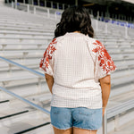 Woman with dark hair standing in a back profile position wearing the Havanah Top at a football field. The focal point of this image is the intricate fabric detail as well as the burnt orange floral embroidery on shoulders. The model paired this top with light-wash denim shorts.