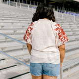 Woman with dark hair standing in a back profile position wearing the Havanah Top at a football field. The focal point of this image is the intricate fabric detail as well as the burnt orange floral embroidery on shoulders. The model paired this top with light-wash denim shorts.