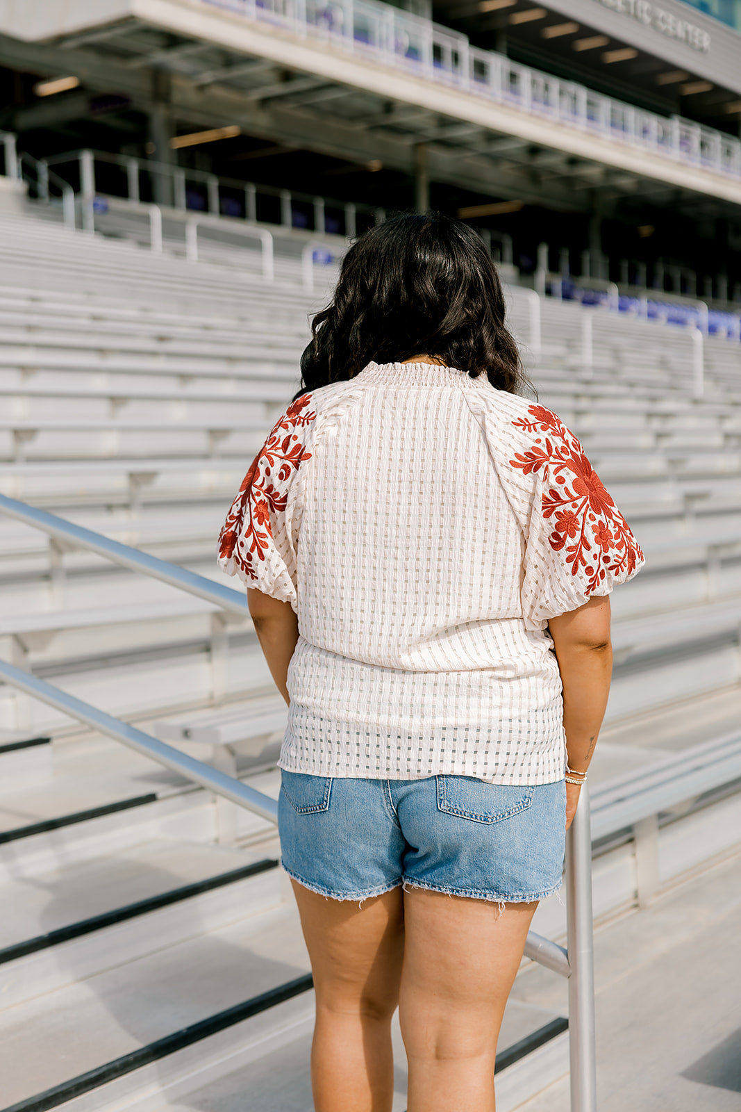 Woman with dark hair standing in a back profile position wearing the Havanah Top at a football field. The focal point of this image is the intricate fabric detail as well as the burnt orange floral embroidery on shoulders. The model paired this top with light-wash denim shorts.