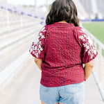 Woman with dark hair standing in a back profile position wearing the Havanah Top at a football field. The focal point of this image is the fabric pattern as well as white floral embroidery down the sleeves. The model paired this top with light-wash denim shorts. 