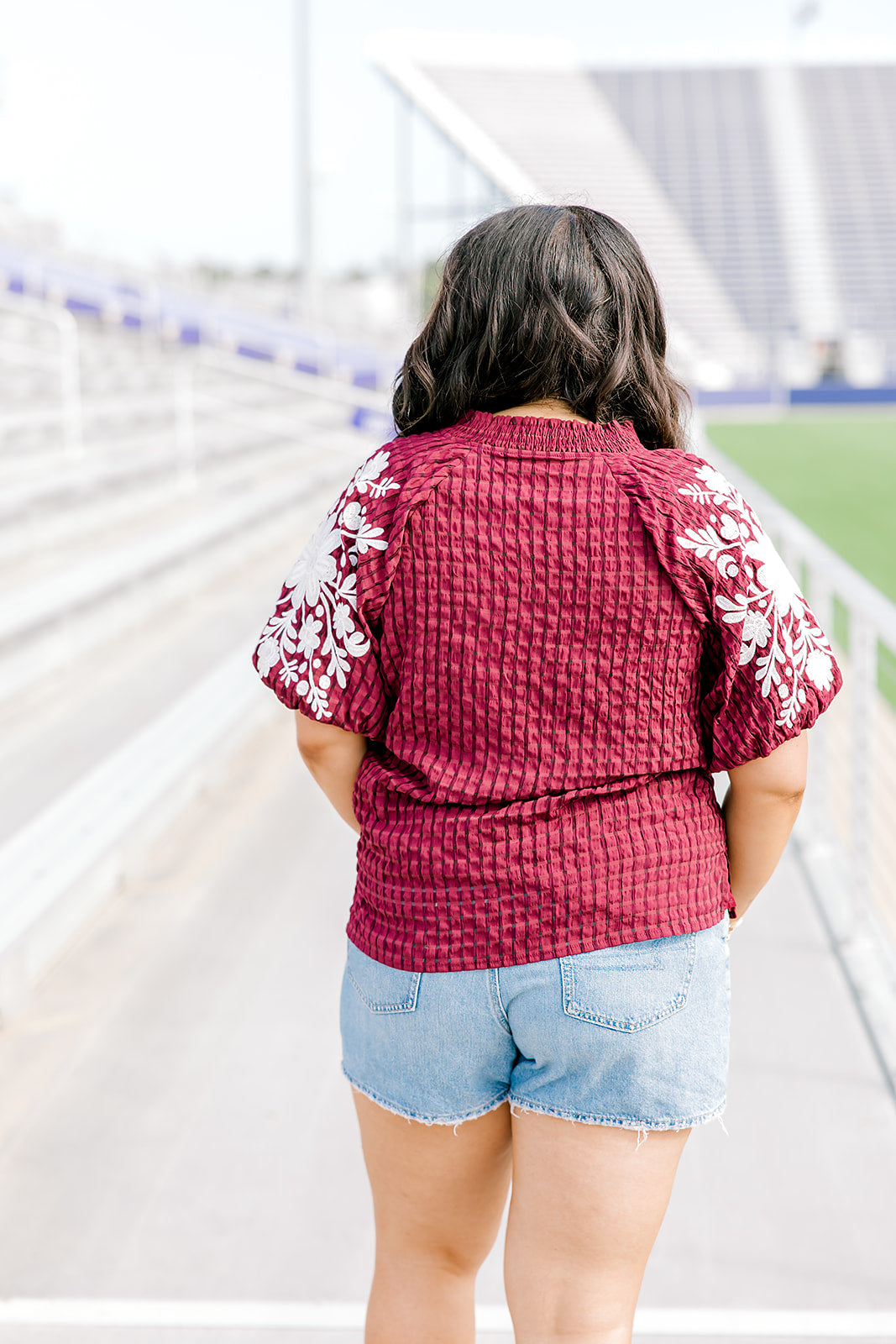 Woman with dark hair standing in a back profile position wearing the Havanah Top at a football field. The focal point of this image is the fabric pattern as well as white floral embroidery down the sleeves. The model paired this top with light-wash denim shorts. 