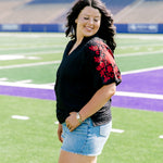 Woman with dark hair wearing a black shirt with red floral embroidery on sleeves. The model paired this top with light-wash denim shorts. 