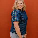 Lady wearing a navy top with colorful embroidery on the sleeve on a orange  background 