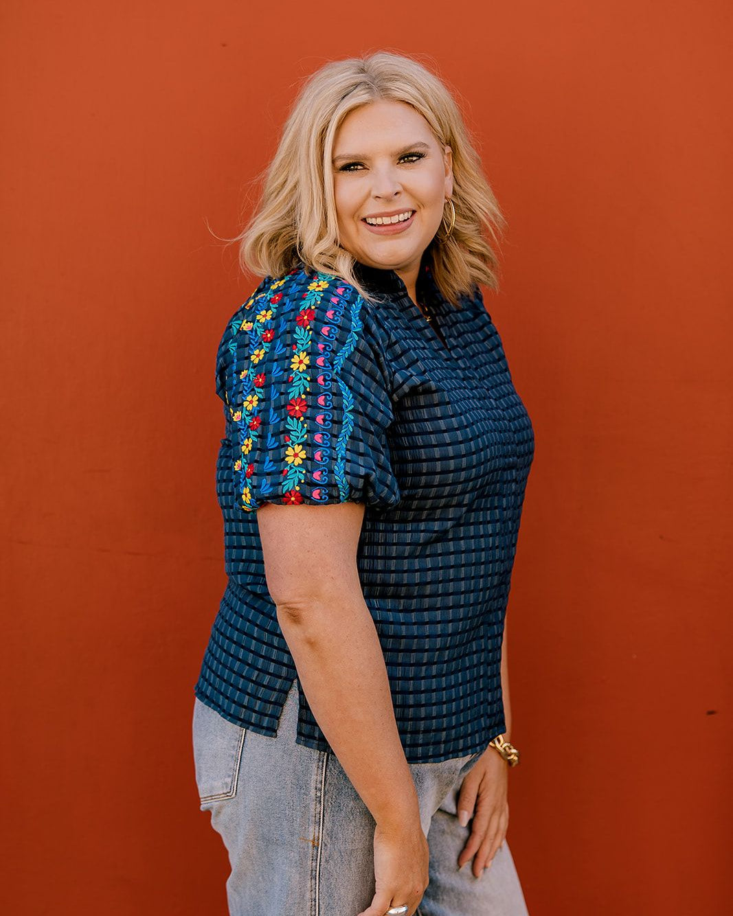Lady wearing a navy top with colorful embroidery on the sleeve on a orange  background 