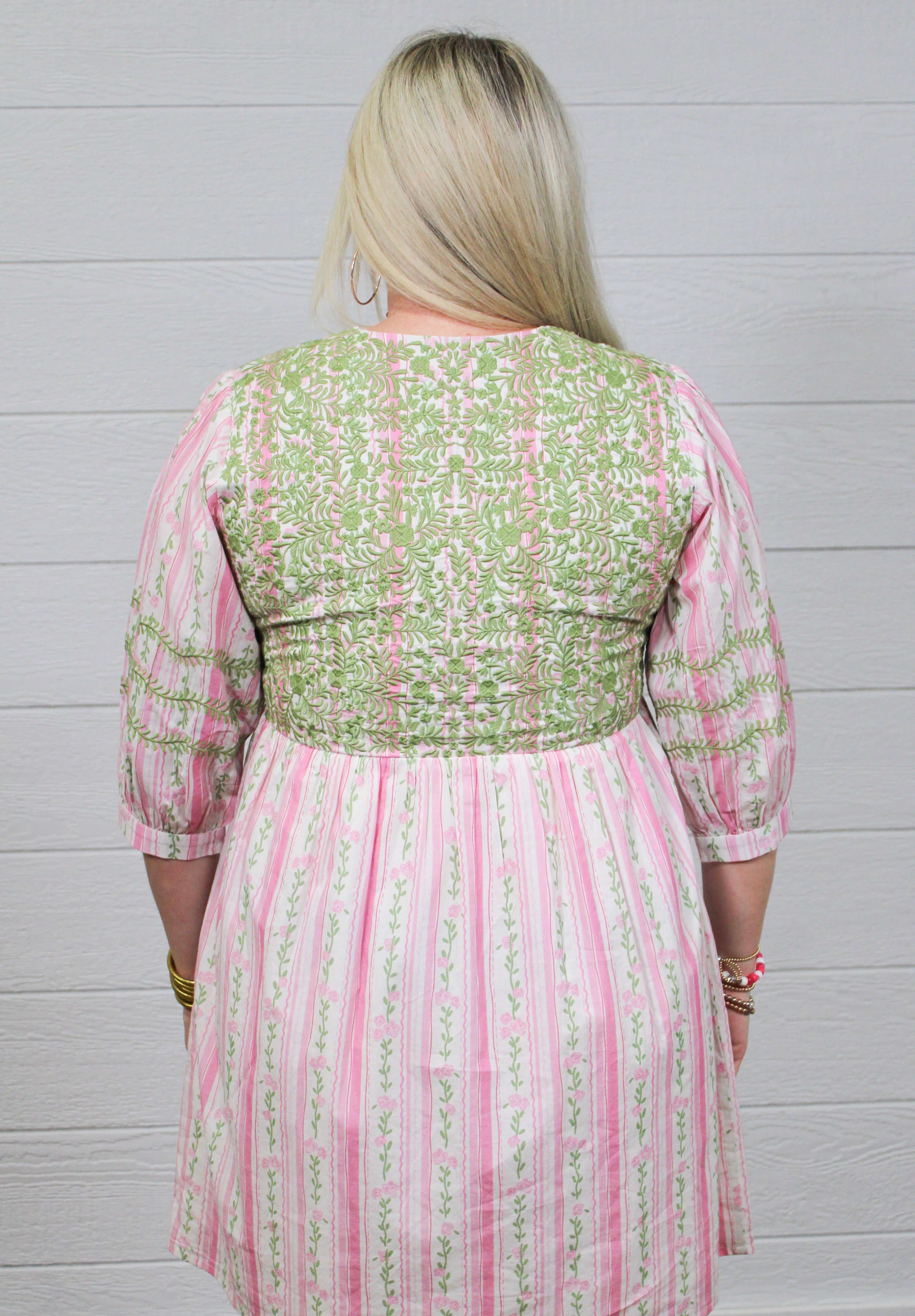 Woman with blonde hair wearing a above-the-knee dress with pink and green floral graphics, and green embroidery on quarter-sleeves, chest, and shoulders. 