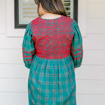 Woman with dark hair wearing a quarter-sleeve dress that has green plaid graphics, red embroidery on back and chest, and on sleeves. 