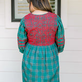 Woman with dark hair wearing a quarter-sleeve dress that has green plaid graphics, red embroidery on back and chest, and on sleeves. 