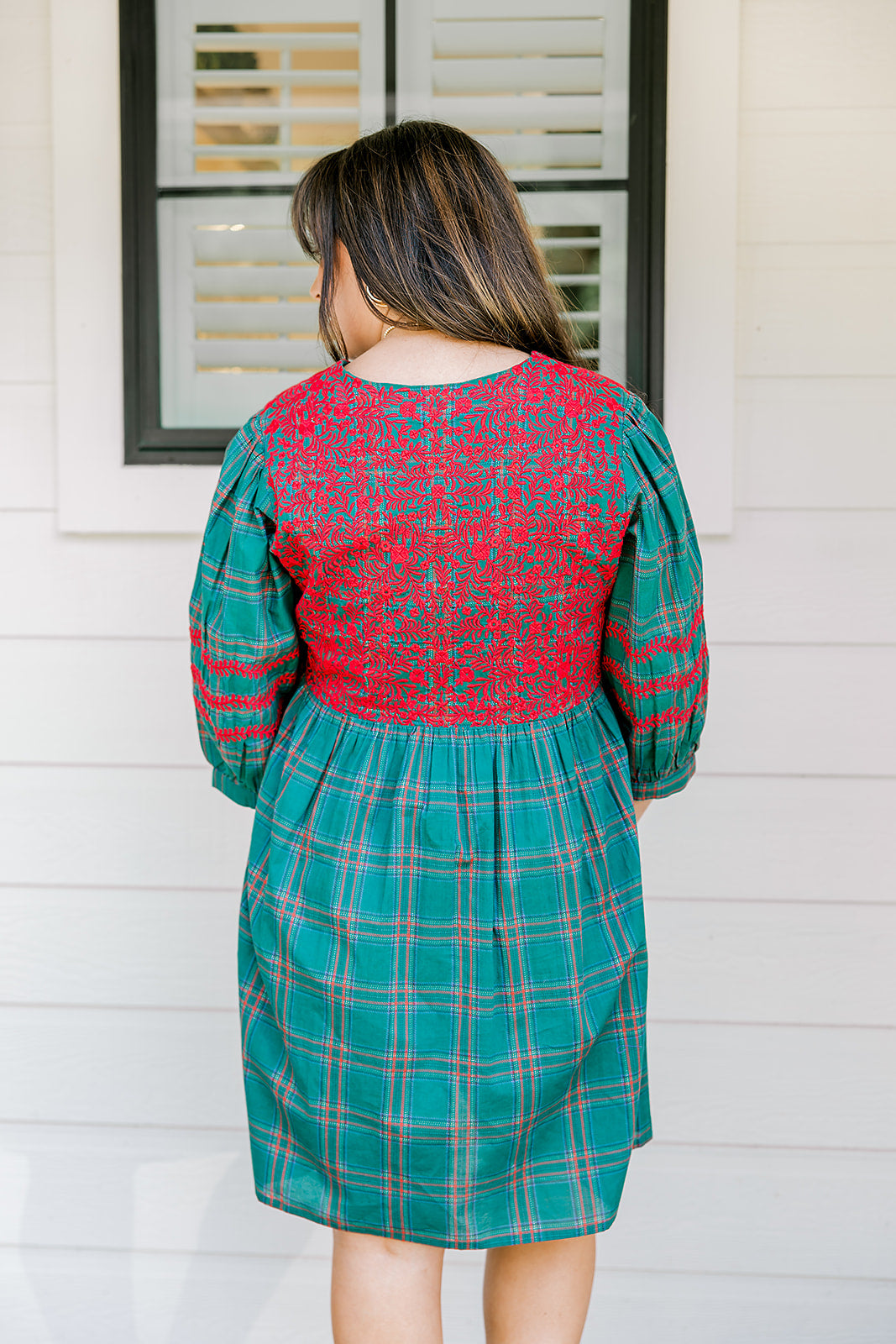 Woman with dark hair wearing a quarter-sleeve dress that has green plaid graphics, red embroidery on back and chest, and on sleeves. 