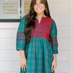 Woman with dark hair wearing a quarter-sleeve dress that has green plaid graphics, red embroidery on back and chest, and on sleeves. 