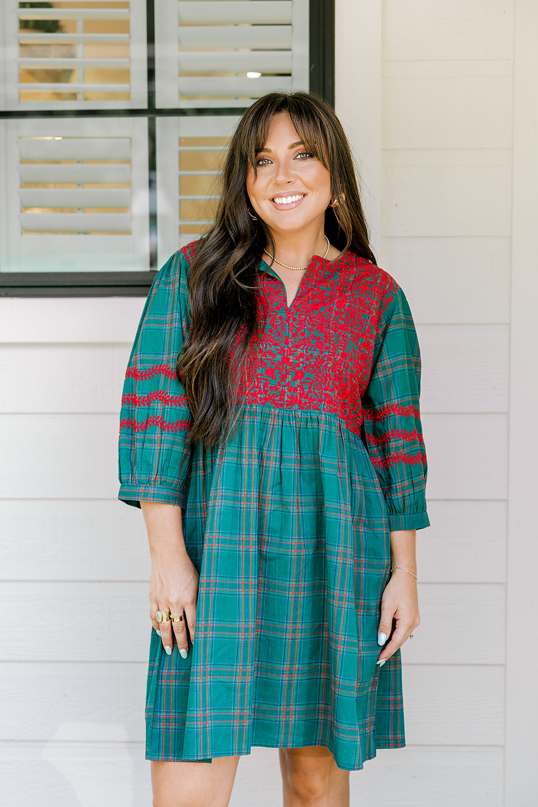 Woman with dark hair wearing a quarter-sleeve dress that has green plaid graphics, red embroidery on back and chest, and on sleeves. 
