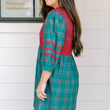 Woman with dark hair wearing a quarter-sleeve dress that has green plaid graphics, red embroidery on back and chest, and on sleeves. 