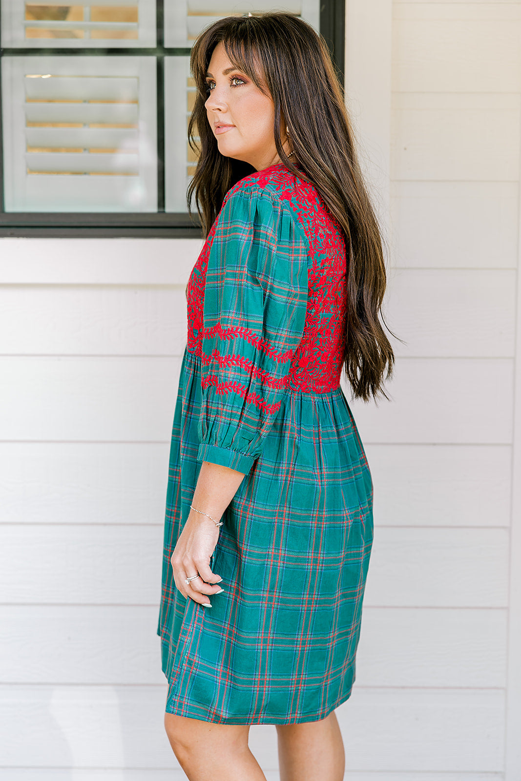 Woman with dark hair wearing a quarter-sleeve dress that has green plaid graphics, red embroidery on back and chest, and on sleeves. 
