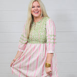 Woman with blonde hair wearing a above-the-knee dress with pink and green floral graphics, and green embroidery on quarter-sleeves, chest, and shoulders. 