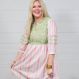 Woman with blonde hair wearing a above-the-knee dress with pink and green floral graphics, and green embroidery on quarter-sleeves, chest, and shoulders. 