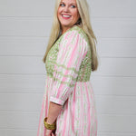 Woman with blonde hair wearing a above-the-knee dress with pink and green floral graphics, and green embroidery on quarter-sleeves, chest, and shoulders. 