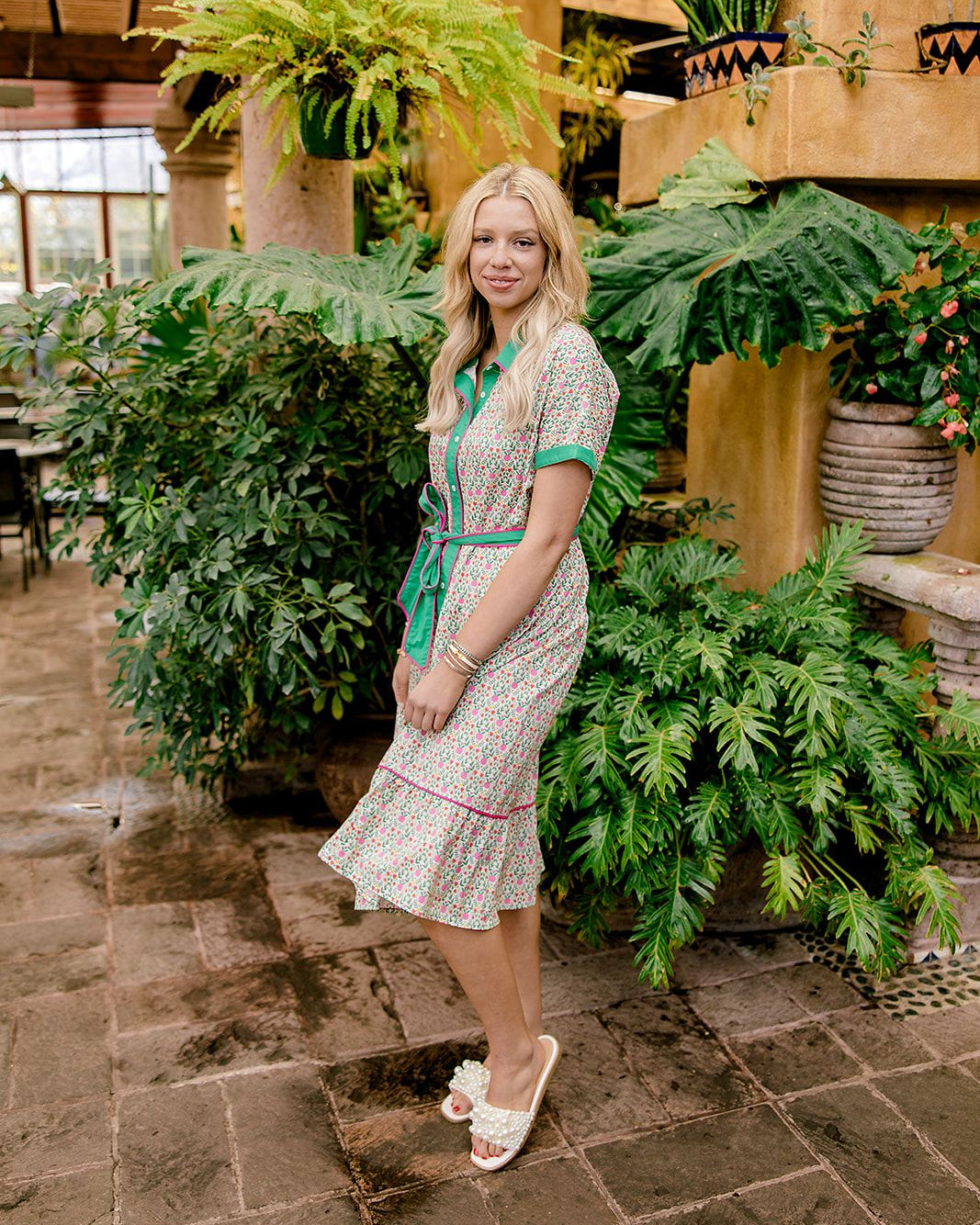 Lady wearing a green and floral dress with pink piping and a tie waist in a leafy natural setting 
