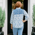 Woman with red hair wearing a quarter-sleeve top with blue Santa toile graphics and blue embroidery on shoulders and chest. Model paired this top with denim jeans.
