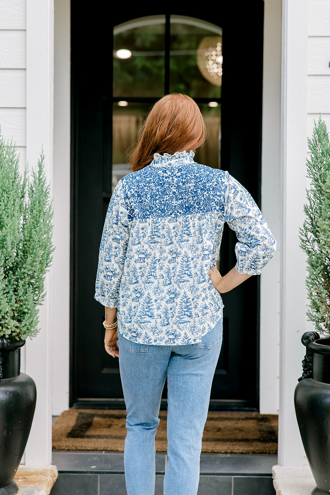Woman with red hair wearing a quarter-sleeve top with blue Santa toile graphics and blue embroidery on shoulders and chest. Model paired this top with denim jeans.