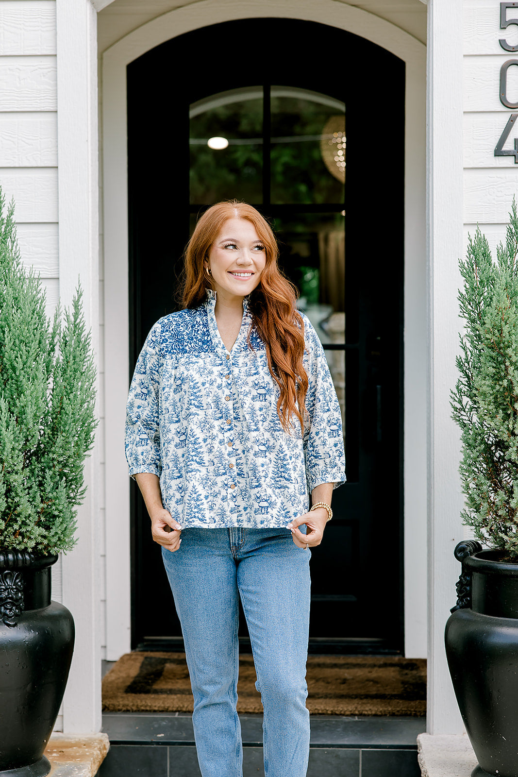 Woman with red hair wearing a quarter-sleeve top with blue Santa toile graphics and blue embroidery on shoulders and chest. Model paired this top with denim jeans.