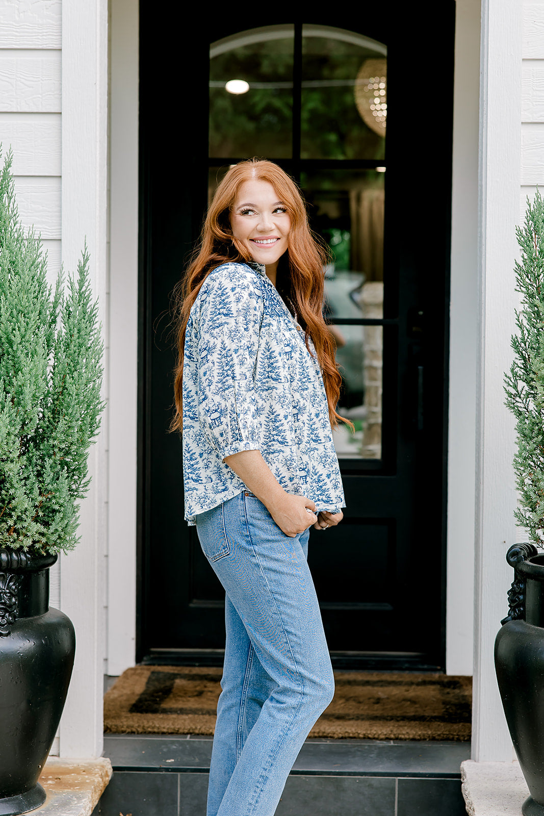 Woman with red hair wearing a quarter-sleeve top with blue Santa toile graphics and blue embroidery on shoulders and chest. Model paired this top with denim jeans.