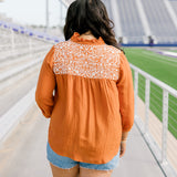 Woman with dark hair standing in a back profile position wearing the Jane Top at a football field. The focal point of this image is the white embroidery across shoulders as well as pleating beneath. The model paired this top with light-wash denim shorts.