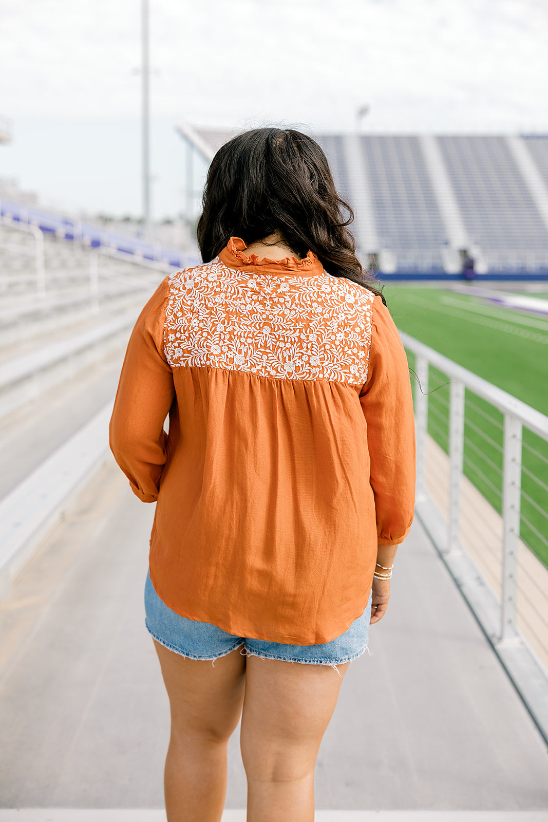 Woman with dark hair standing in a back profile position wearing the Jane Top at a football field. The focal point of this image is the white embroidery across shoulders as well as pleating beneath. The model paired this top with light-wash denim shorts.