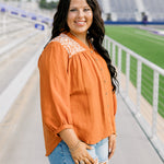 Woman with dark hair standing in a side profile position wearing the Jane Top at a football field. The focal point of this image is the quarter sleeve with pleating around the cuff as well as white embroidery along shoulders. The model paired this top with light-wash denim shorts.