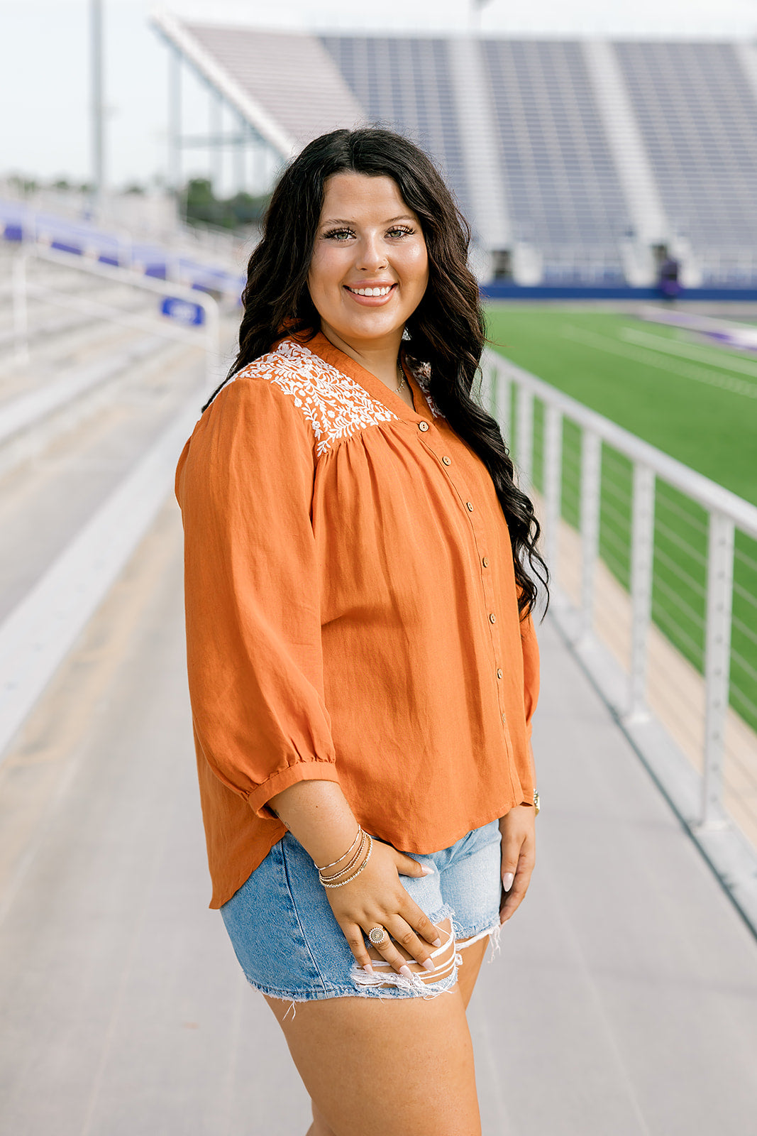 Woman with dark hair standing in a side profile position wearing the Jane Top at a football field. The focal point of this image is the quarter sleeve with pleating around the cuff as well as white embroidery along shoulders. The model paired this top with light-wash denim shorts.