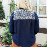 Woman with blonde hair wearing blue quarter sleeve blouse with white embroidery on front and back. and a burnt orange skirt.