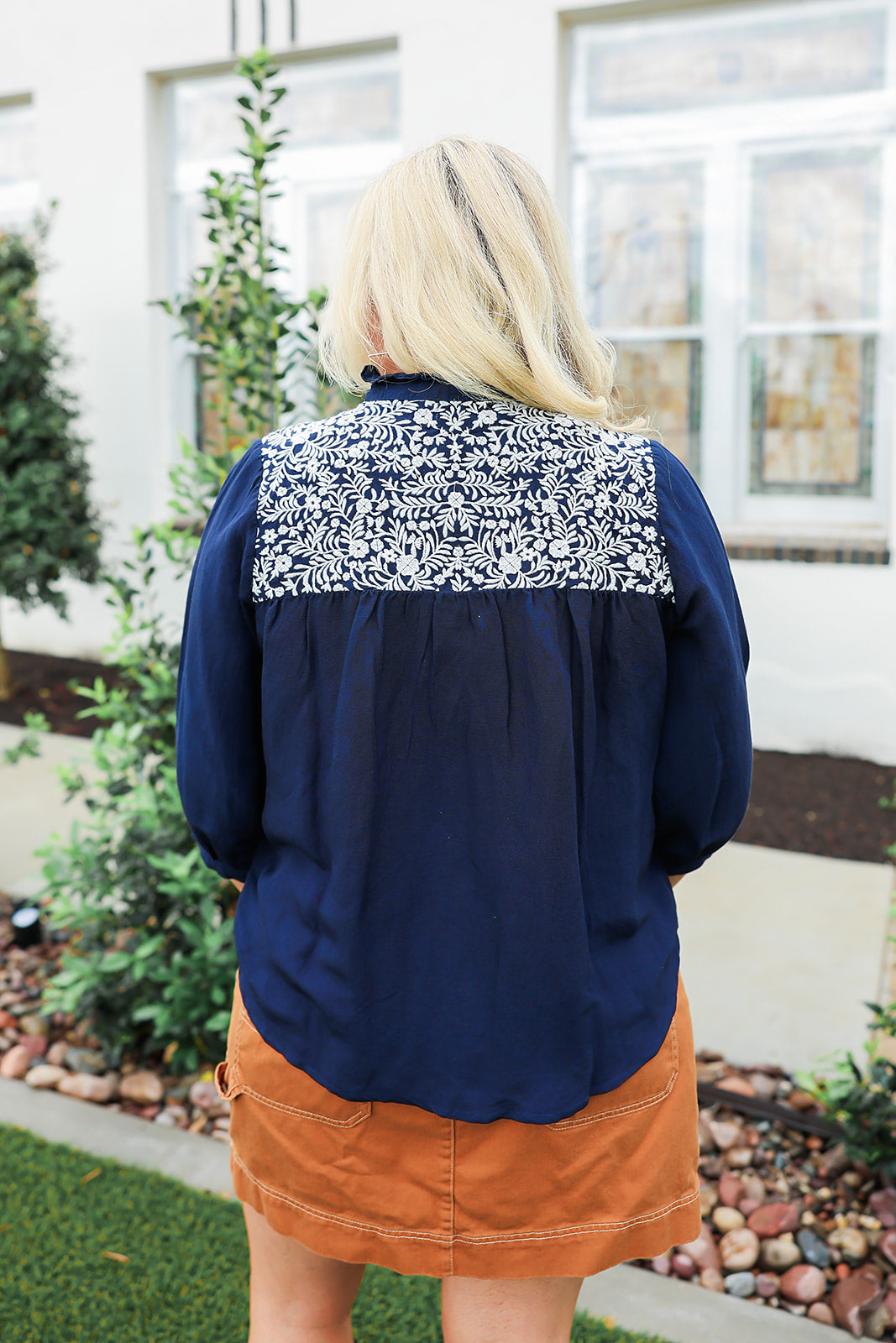Woman with blonde hair wearing blue quarter sleeve blouse with white embroidery on front and back. and a burnt orange skirt.