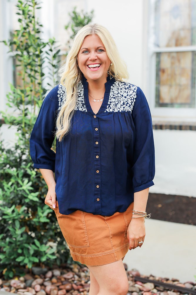 Woman with blonde hair wearing blue quarter sleeve blouse with white embroidery on front and back. and a burnt orange skirt.