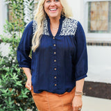 Woman with blonde hair wearing blue quarter sleeve blouse with white embroidery on front and back. and a burnt orange skirt.