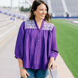 Woman with dark hair standing in a front profile position wearing the Jane Top at a football field. The focal point of this image is the embroidery on shoulders, buttons down the front of top, high-collar and v-neckline. The model paired this top with dark-wash denim shorts.