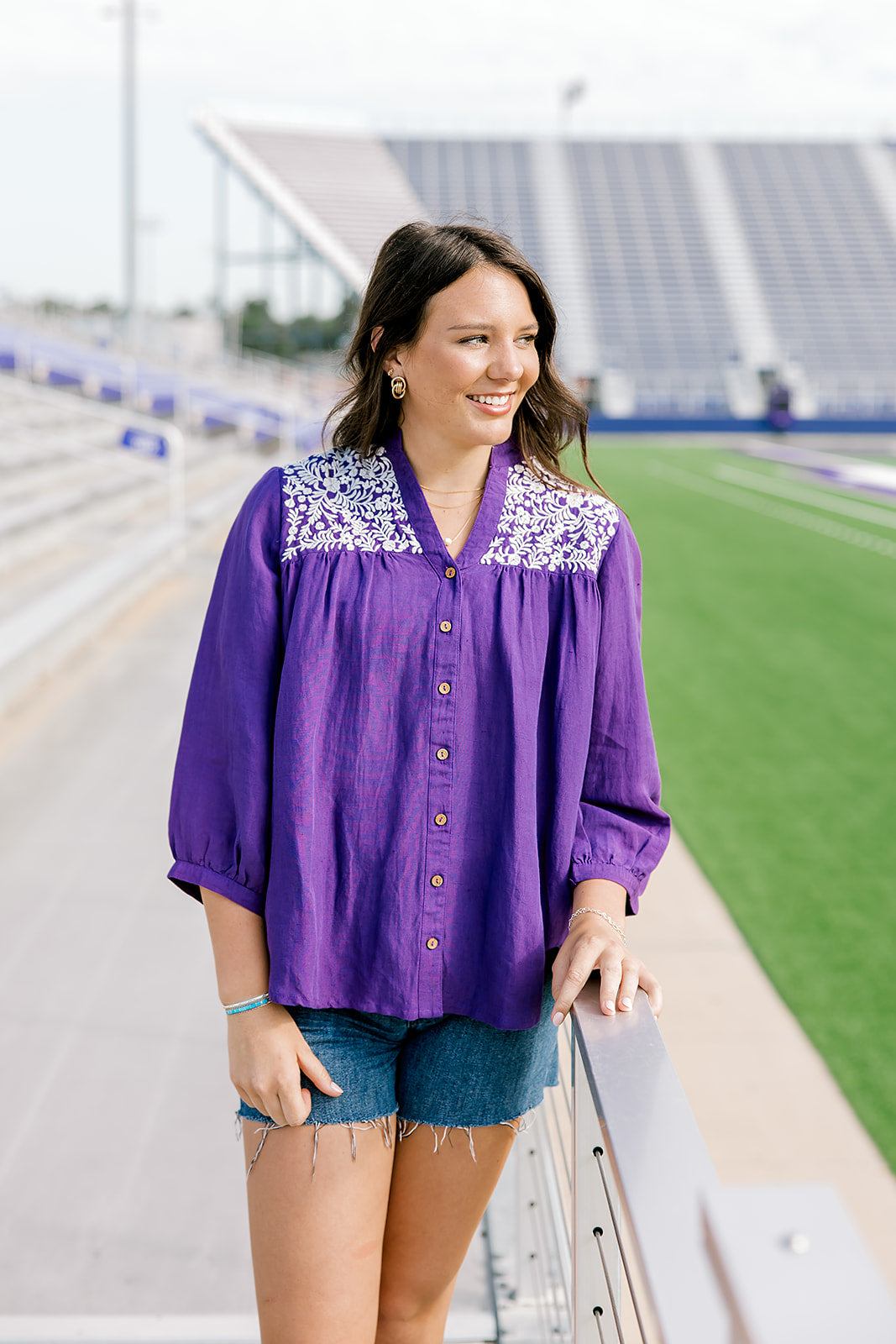 Woman with dark hair standing in a front profile position wearing the Jane Top at a football field. The focal point of this image is the embroidery on shoulders, buttons down the front of top, high-collar and v-neckline. The model paired this top with dark-wash denim shorts.