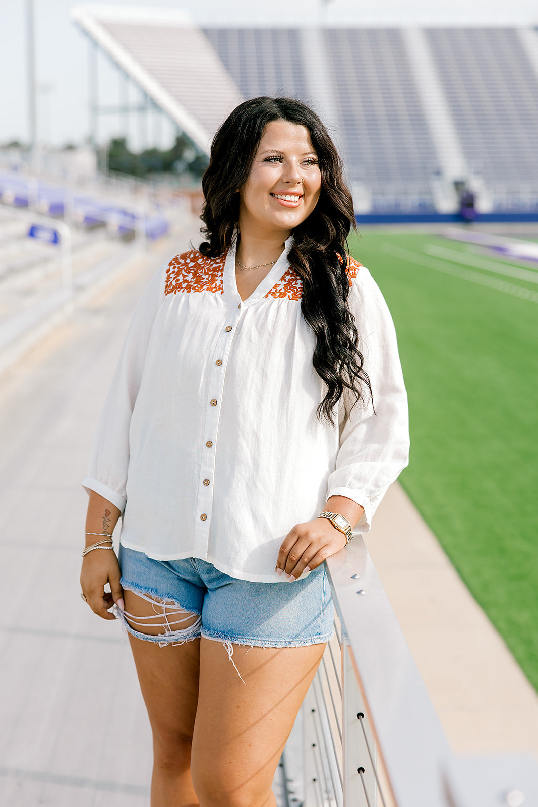 Woman with dark hair standing in a front profile position wearing the Jane Top at a football field. The focal point of this image is the buttons down the front of the top, the burnt orange embroidery as well as the v-neckline. The model paired this top with light-wash denim shorts.