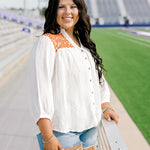 Woman with dark hair standing in a side profile position wearing the Jane Top at a football field. The focal point of this image is the buttons down the front of top as well as burnt orange embroidery on shoulders. The model paired this top with light-wash denim shorts.