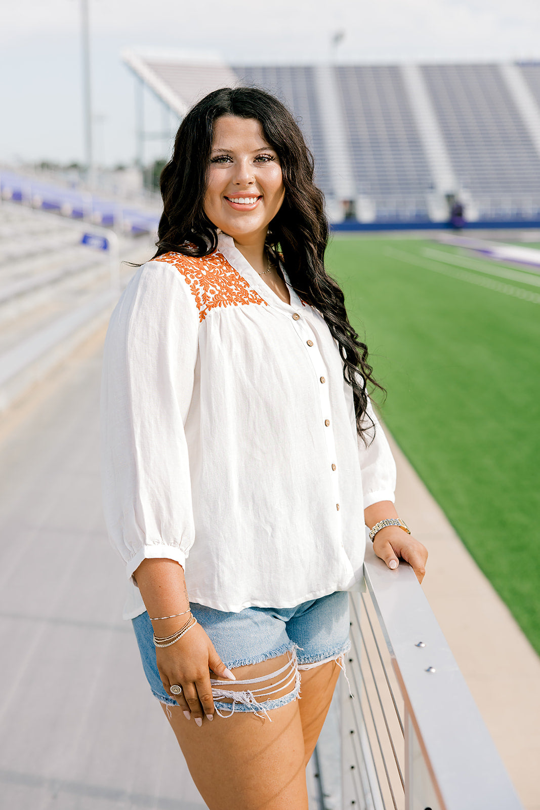 Woman with dark hair standing in a side profile position wearing the Jane Top at a football field. The focal point of this image is the buttons down the front of top as well as burnt orange embroidery on shoulders. The model paired this top with light-wash denim shorts.