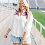 Woman with blonde hair standing in a front profile position wearing the Jane Top at a football field. The focal point of this image is the embroidery across front of top, buttons down the front of shirt and the v-neckline with a high collar. The model paired this top with light-wash denim shorts.