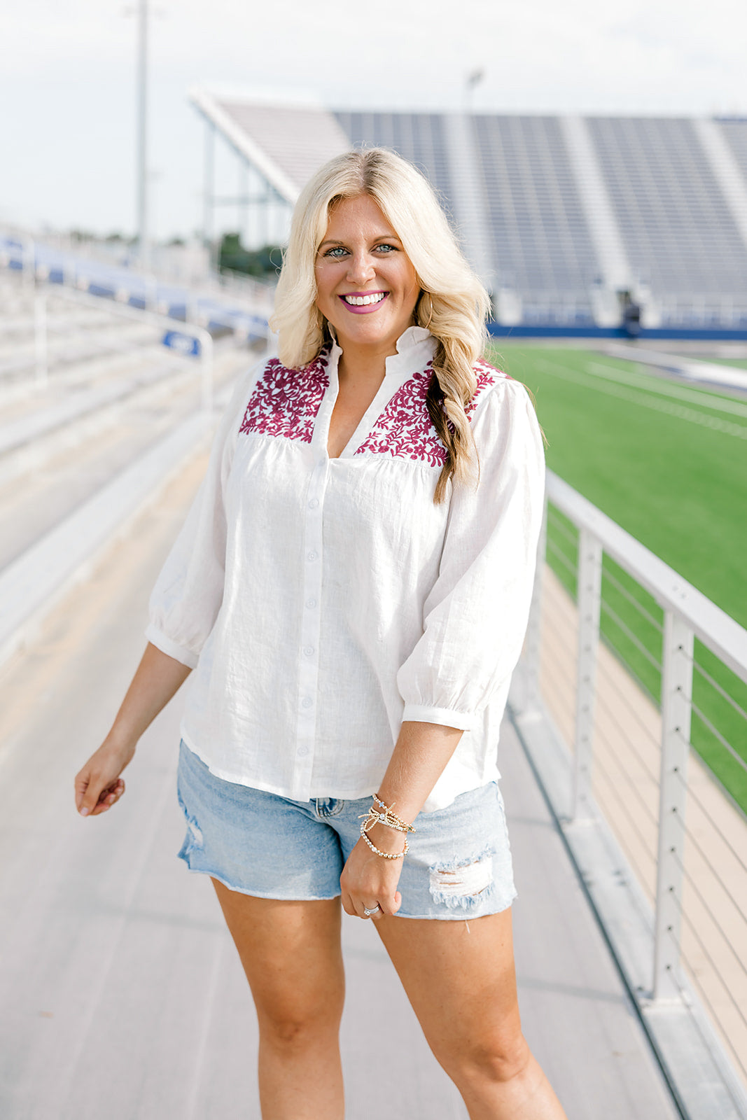 Woman with blonde hair standing in a front profile position wearing the Jane Top at a football field. The focal point of this image is the embroidery across front of top, buttons down the front of shirt and the v-neckline with a high collar. The model paired this top with light-wash denim shorts.