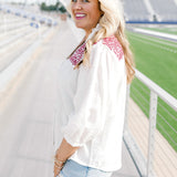 Woman with blonde hair standing in a side profile position wearing the Jane Top at a football field. The focal point of the top is the quarter sleeve length as well as pleating towards cuff. There is also an emphasis on the embroidery across the shoulders of top. The model paired this top with light-wash denim shorts.