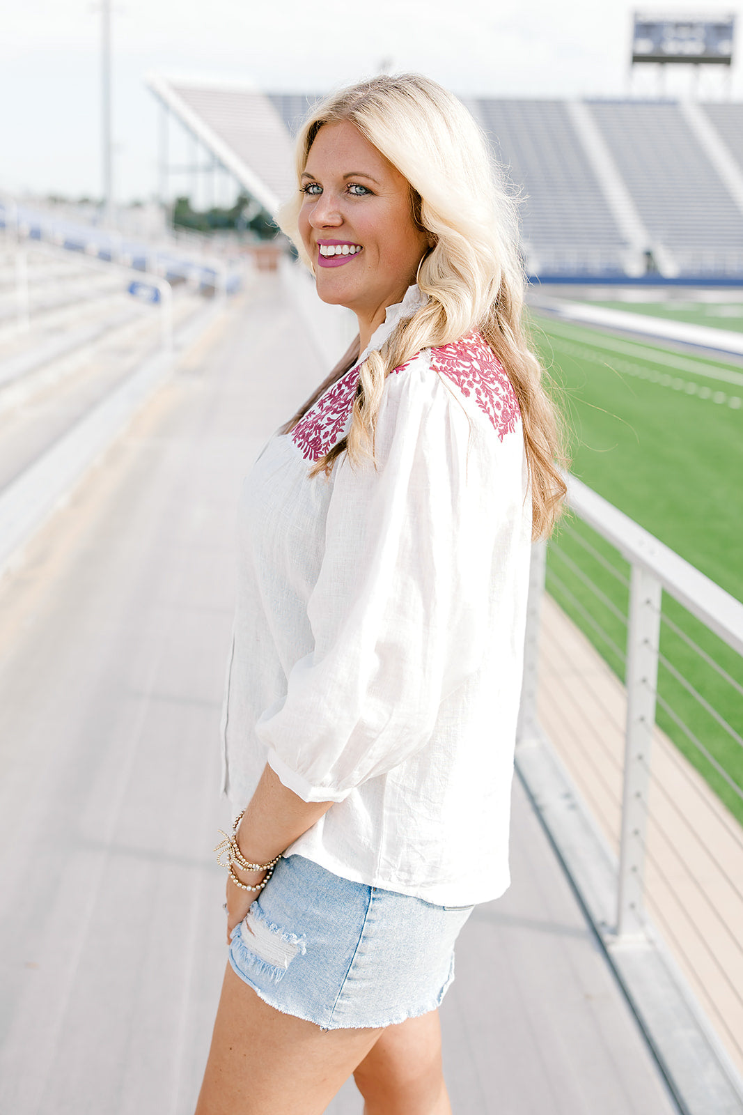 Woman with blonde hair standing in a side profile position wearing the Jane Top at a football field. The focal point of the top is the quarter sleeve length as well as pleating towards cuff. There is also an emphasis on the embroidery across the shoulders of top. The model paired this top with light-wash denim shorts.