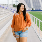 Woman with dark hair standing in a front profile position wearing the Jane Top at a football field. The focal point of this image is the buttons down the front of the top as well as the embroidery down shoulders and v-neckline. The model paired this top with light-wash denim shorts.