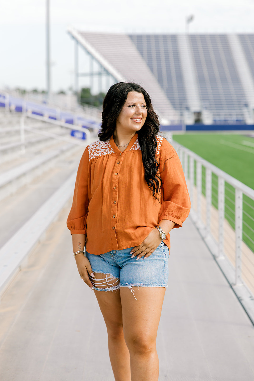 Woman with dark hair standing in a front profile position wearing the Jane Top at a football field. The focal point of this image is the buttons down the front of the top as well as the embroidery down shoulders and v-neckline. The model paired this top with light-wash denim shorts.