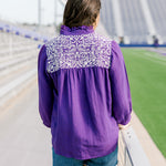 Woman with dark hair standing back profile wearing the Jane Top at a football field. The focal point of this image is the embroidery along the shoulders, pleated back, and high collar. The model paired this top with dark-wash denim shorts.