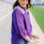 Woman with dark hair standing in a side profile position wearing the Jane Top at a football field. The focal point of this image is the quarter sleeve pleated sleeves cuffed at bottom, embroidery along the shoulders, and a sneak peak at the buttons down the front. The model paired this top with dark-wash denim shorts.