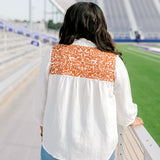 Woman with dark hair standing in a back profile position wearing the Jane Top at a football field. The focal point of this image is the pleating beneath embroidery and the burnt orange embroidery across shoulders. The model paired this top with light-wash denim shorts.